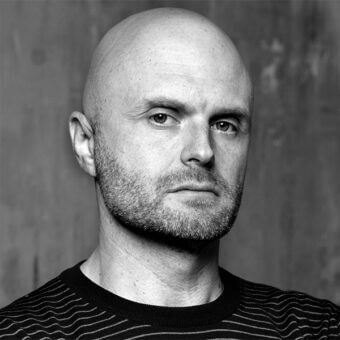 Black and white portrait of Robin Rizzini, a man with shaved head, salt-and-pepper beard, wearing a dark striped sweater, against a mottled gray wall.