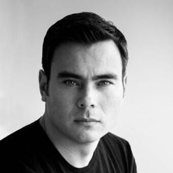 Monochromatic portrait of a man facing forward with dark hair, neutral expression, and wearing a dark, smooth garment against a light gray background
