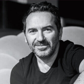 Sacha Lakic portrait, man with dark hair and beard seated against a white rounded upholstered lounge chair, neutral expression.