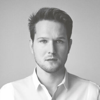 Sebastian Herkner portrait, grayscale photograph, man with beard wearing white shirt, neutral expression, gradient gray background.