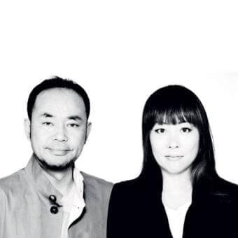 Two people face forward against a light backdrop in grayscale, wearing jackets and collared shirts with neutral expressions
