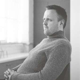 Black and white portrait of Simon Pengelly in grey knit turtleneck, seated in profile, gazing out a window with metal framing.