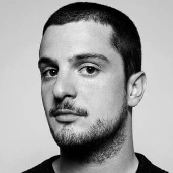 Close-up black and white portrait of a young man facing forward with short hair and a partially grown beard against a light gray background