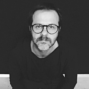 Portrait of Simone Fanciullacci, a man with salt-and-pepper beard wearing black glasses and t-shirt, against a dark background.