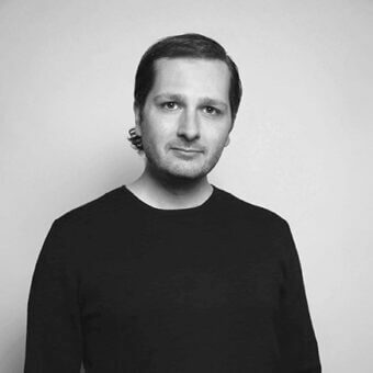 Portrait of Stefan Krivokapic, dark sweater, short brown hair, silver earring, neutral expression, light gray background, studio lighting.