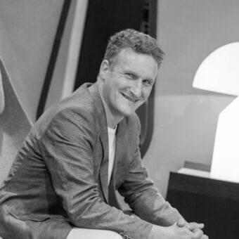 Man with curly hair smiles while seated wearing a collared shirt and textured jacket near a curved lamp
