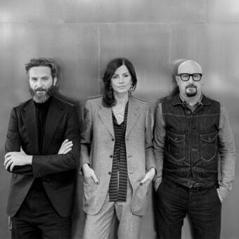 Three people in tailored clothing stand against a vertically paneled grayscale background with direct forward gazes