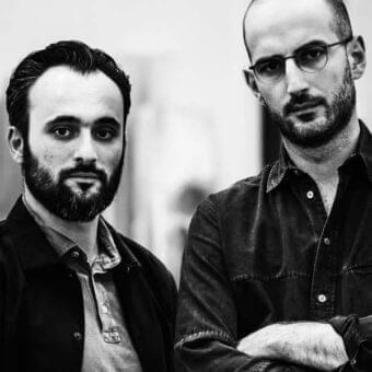 Two men with dark hair and facial hair are shown in grayscale, facing forward with crossed arms against a light background