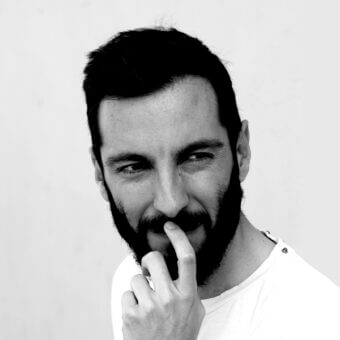 Black and white portrait of Tiziano Guardini, light gray background, short dark hair, beard, white t-shirt, soft lighting, close-up composition.