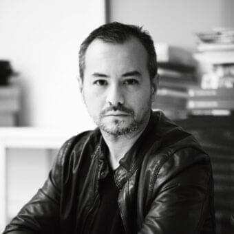 Grayscale portrait of a man with short dark hair and beard wearing a dark jacket, facing forward against a blurred shelving unit