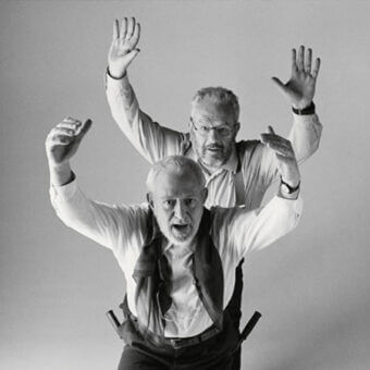 Black and white studio portrait of two men in white shirts, one with a vest and rifles, displaying dramatic expressions and raised arms.