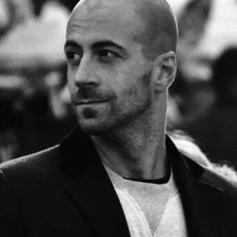 Grayscale portrait of a shaved-headed man with stubble wearing a light textured top and dark jacket looking slightly right