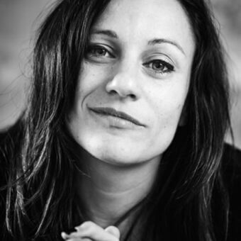 Black and white portrait of Viola Tonucci, centered face, dark wavy hair, soft lighting, neutral blurred background, dark knit garment.