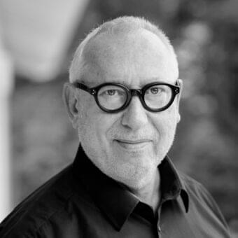 Grayscale portrait of an older person with gray beard and round glasses wearing a dark collared shirt against a blurred outdoor background
