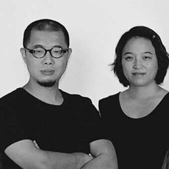 Black and white studio portrait of two people, a man with goatee and a woman with bob haircut, both in black clothing against a light backdrop.