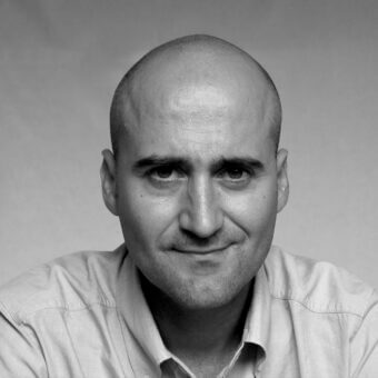 Black and white portrait of Alessandro Piva, a bald man with a light shirt, facing forward with a slight smile against a gradient gray background.