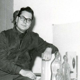 Angelo Barovier portrait, man in black turtleneck, seated with glass bottles, white object, and wedding band on hand, interior setting.