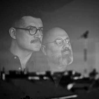 Layered portrait of two men with glasses, one bald with beard, other with dark wavy hair, against blurred urban backdrop, watermark text "calvi brambilla".
