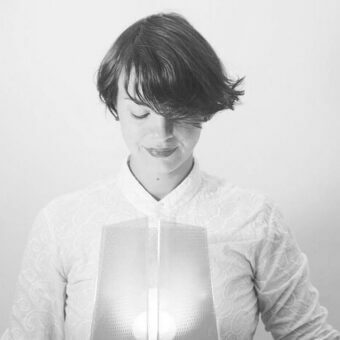 Woman in floral shirt with brushed aluminum panel, layered dark hair, closed eyes, soft lighting, minimalist gray background.