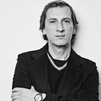 Philippe Nigro portrait, man with brown hair, black blazer, crossed arms, silver wristwatch, light gray background, minimalist aesthetic.