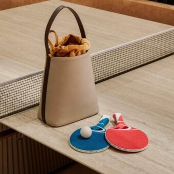 Table tennis table, beige laminate, with white net and metal posts, featuring a tan leather handbag, blue/red paddles, and a white ball.