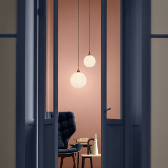 Tee suspension lamp, bronze & white glass, spherical shades illuminate terracotta wall, navy blue doorway, wingback chair visible.