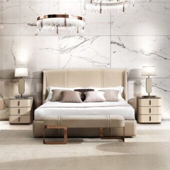 King bed with beige upholstered headboard, rose gold bench, dark grey & beige nightstands, acrylic chandelier, and marble tile backdrop.