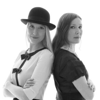 Two women, black and white portrait, arms crossed, facing forward, wearing contrasting tops and hats against a plain backdrop.