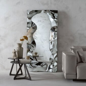 Rectangular mirror with fragmented glass and chrome frame, beside a dark coffee table with vase, sculpture, and books, near a gray sofa.