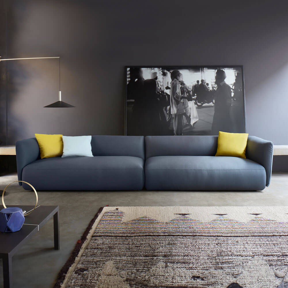 Gray three-seater sofa with textured fabric, mustard & gray pillows, low profile, on patterned rug in a contemporary room setting.