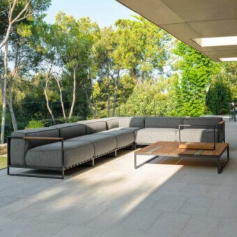Modular sectional sofa, dark gray fabric, square tufting, with matching wood and metal coffee table on gray tiled patio, shaded by beige awning.