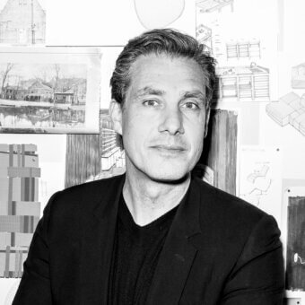 Black and white portrait of Stefan Scholten amidst architectural drawings, featuring sketches of houses, furniture, and geometric designs pinned to a wall.