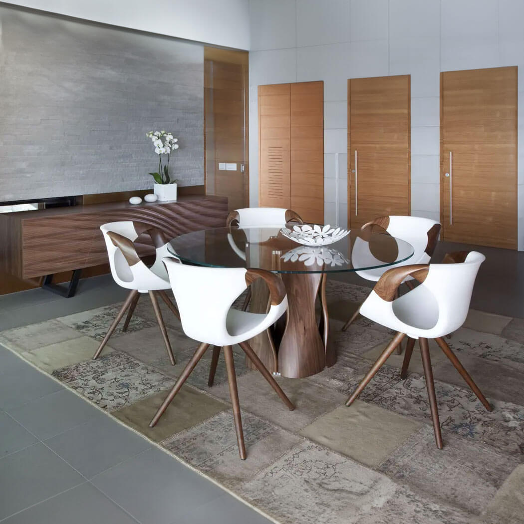 Dining room with round glass table, walnut pedestal, six white molded plastic chairs, gray tile floor, and wood sideboard against a gray wall.