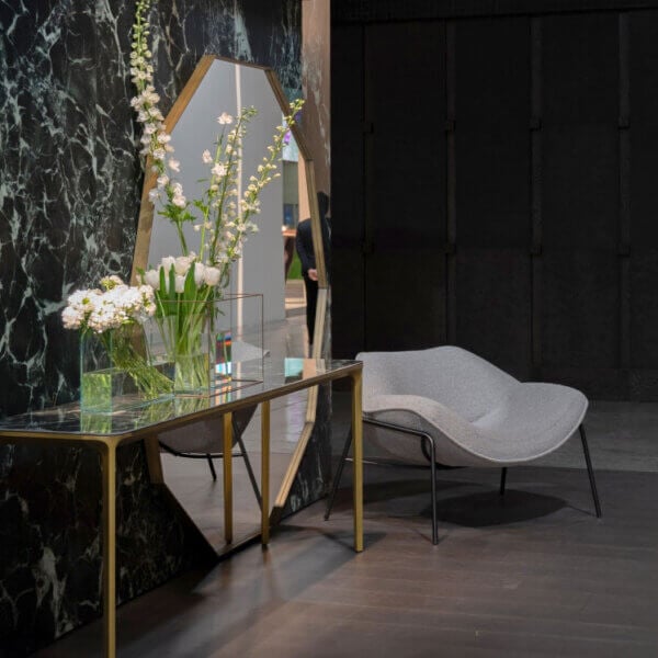 Marble console table with gold legs, black top, and glass case, beside a gray chaise lounge in a marbled room.