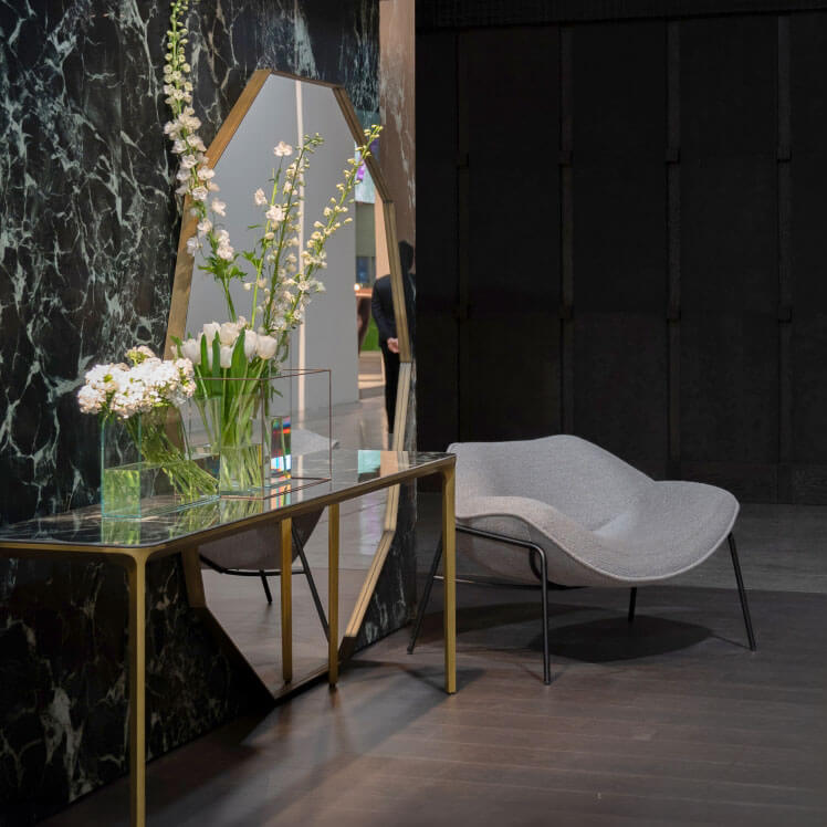Marble console table with gold legs, black top, and glass case, beside a gray chaise lounge in a marbled room.