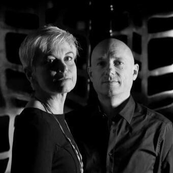 Black and white portrait of a woman with white hair and a bald man, against a geometric textured wall with rectangular patterns.