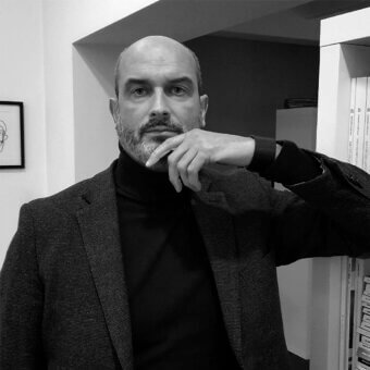 Maurizio Manzoni portrait, black turtleneck and gray blazer, leaning against shelving unit with architectural documents in studio setting.