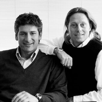 Two men in sweaters and collared shirts seated indoors, dark and light tones, horizontal blinds background.