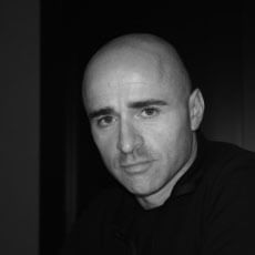 Black and white portrait of Alessandro Elli, a bald man with a neutral expression, wearing a dark crew-neck shirt against a dark background.