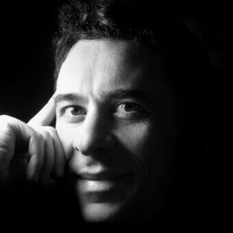 Black and white portrait of Angelo Tomaiuolo, three-quarter view, hand near temple, dark curly hair, dramatic chiaroscuro lighting, studio shot.