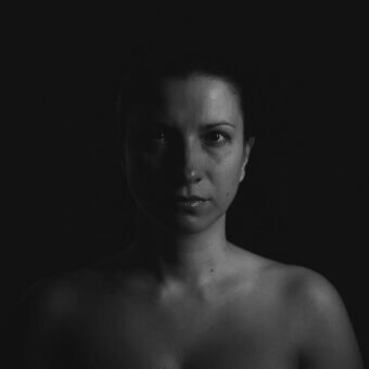 Black and white portrait of a woman with direct eye contact, smooth skin, and bare shoulders against a dark background.