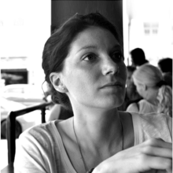 Black and white portrait of Beatrice Fanchini, dark hair in a bun, light t-shirt, contemplative gaze, blurred interior background, soft lighting.
