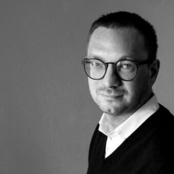 Black and white portrait of Cesare Arosio, a man with dark hair wearing glasses and a white shirt under a dark v-neck sweater, neutral gray background.