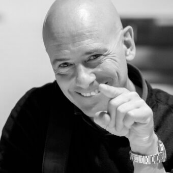 Black and white portrait of Daniele Lo Scalzo Moscheri, a smiling bald man in a black textured sweater, wearing a silver wristwatch.