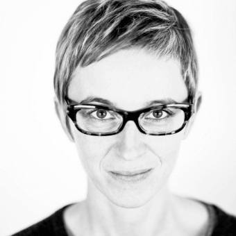Black and white portrait of a woman with short hair, wearing dark eyeglasses, neutral expression, studio lighting, white background.