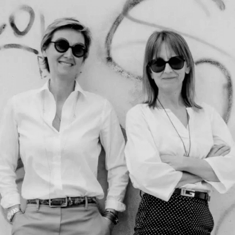 Black and white portrait of two women in shirts and trousers/skirt, against a textured wall with abstract graffiti markings and even lighting.