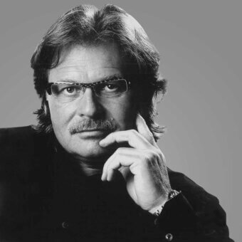 Man in glasses, black turtleneck, and jacket, with gray hair and mustache, hand to cheek, silver wristwatch, gray gradient background.