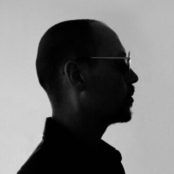 Bald man portrait, grayscale, rectangular glasses, facing left, dark shirt, short beard, gradient background, studio lighting, silhouette effect.