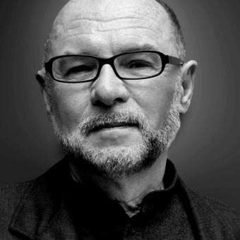 Portrait of Peter Maly, a middle-aged man with a shaved head and gray beard, wearing glasses and a dark mandarin collar shirt.