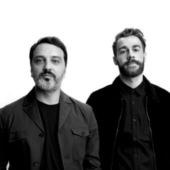 Black and white portrait of two men in dark blazers, minimalist studio shot with even lighting, focused expressions, white background.